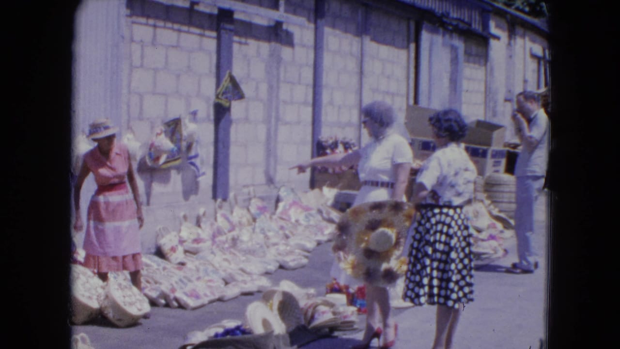 In a 1964 Kingston market scene, women in dresses look at woven baskets and broad-brim hats laid out in bright sun.