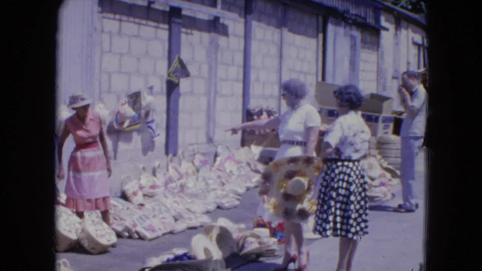 In a 1964 Kingston market scene, women in dresses look at woven baskets and broad-brim hats laid out in bright sun.