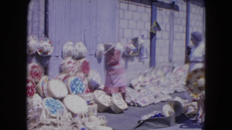 A woman in a pink dress stands amid colorful woven hats and baskets in the Kingston market footage.