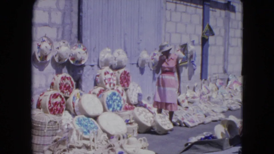 A woman in a pink dress holds a broad-brim hat beside piles of baskets and colorful woven hats in Kingston.