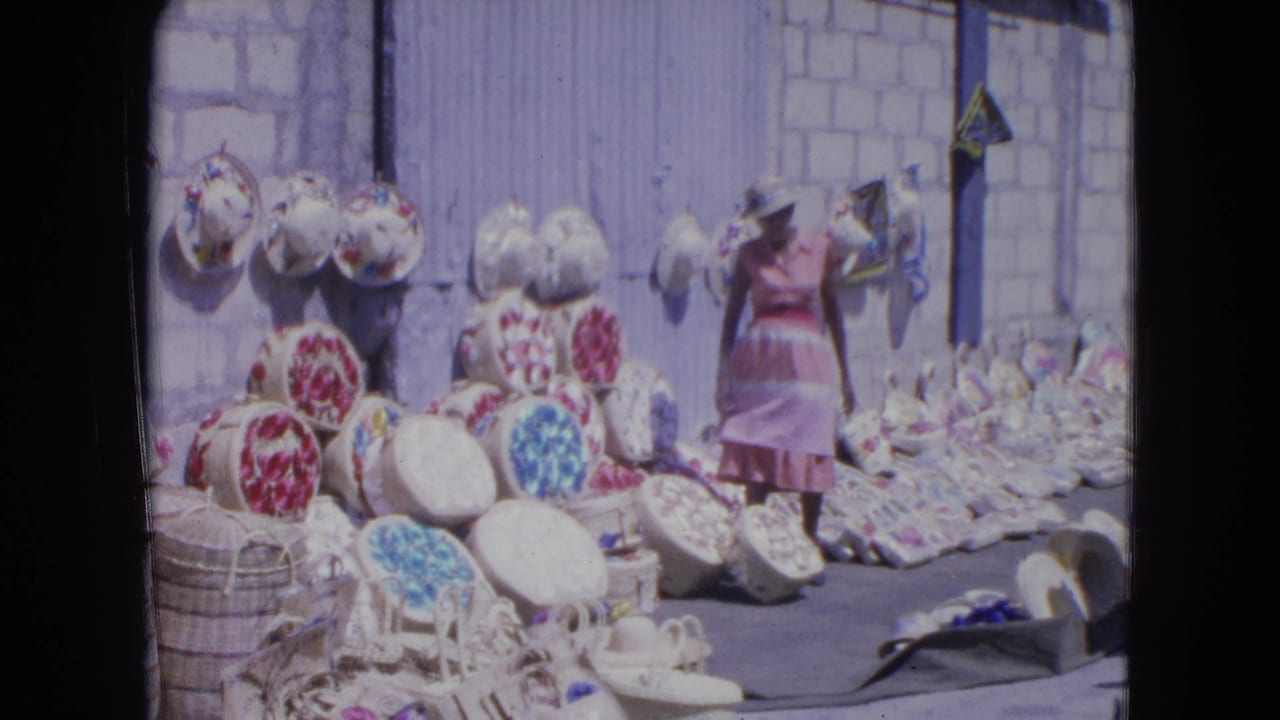 In Kingston, Jamaica in 1964, a woman stands near handwoven straw hats and baskets arranged on the ground and wall.