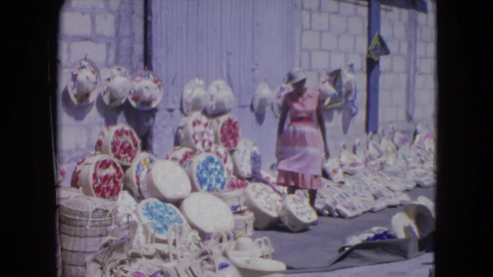 In Kingston, Jamaica in 1964, a woman stands near handwoven straw hats and baskets arranged on the ground and wall.