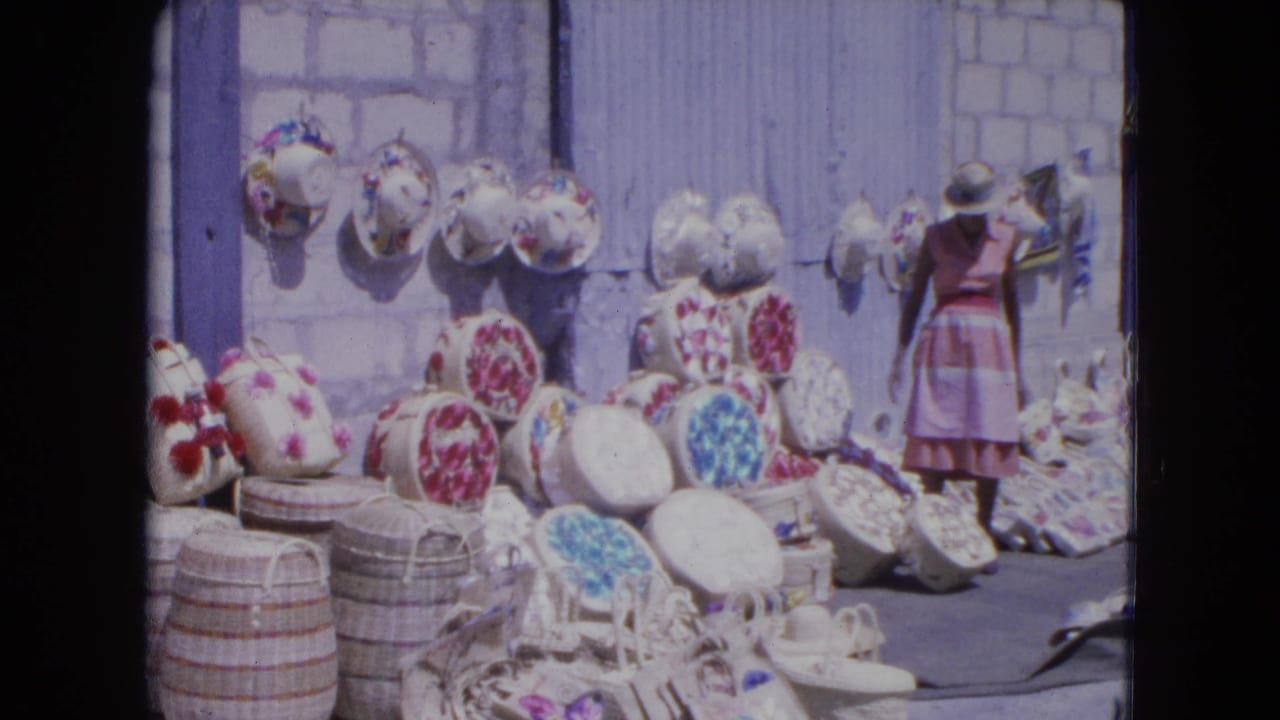 A 1964 Kingston market scene shows a woman beside colorful straw hats and baskets displayed outdoors against masonry.
