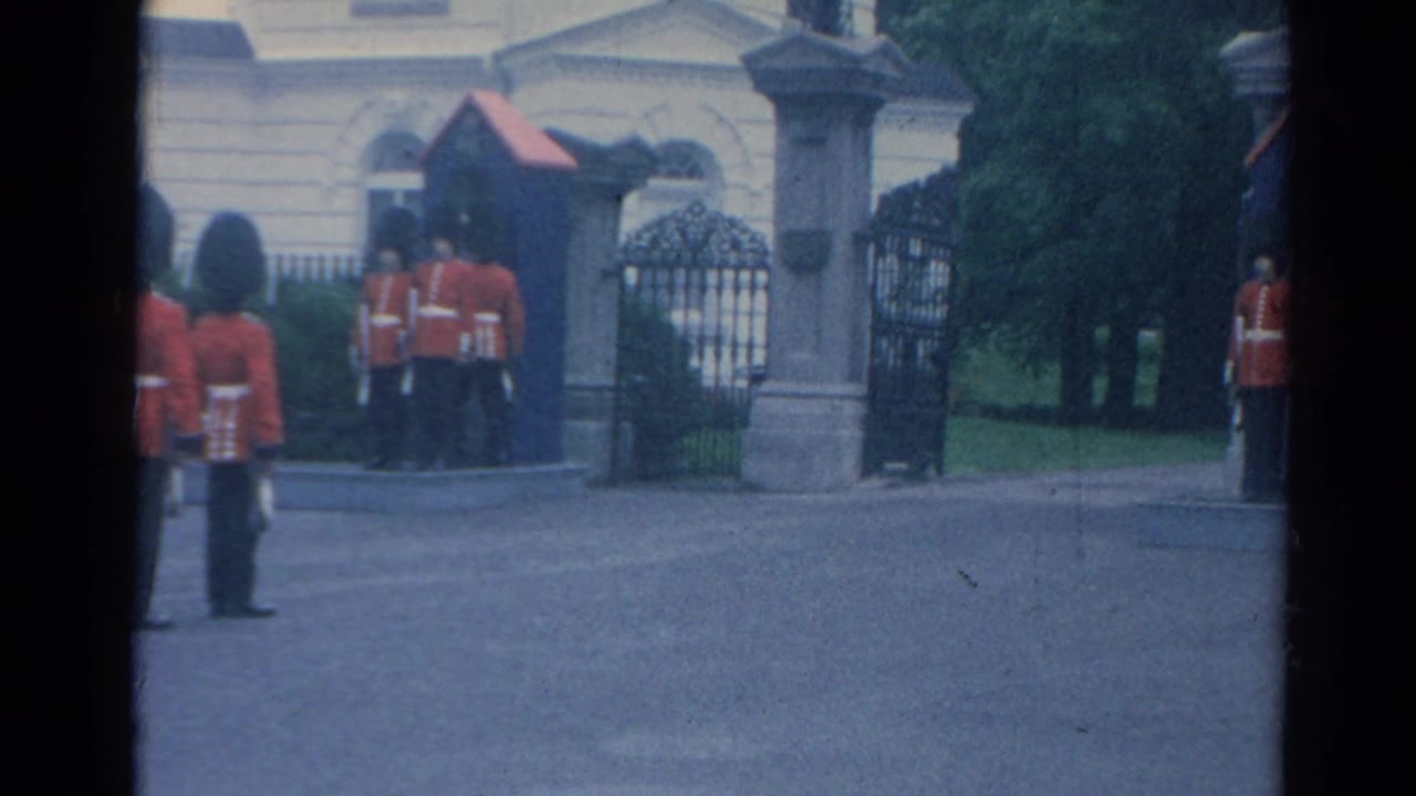1964 London home-movie frame showing ceremonial guards near the Buckingham Palace entrance, with gates and stonework behind them.