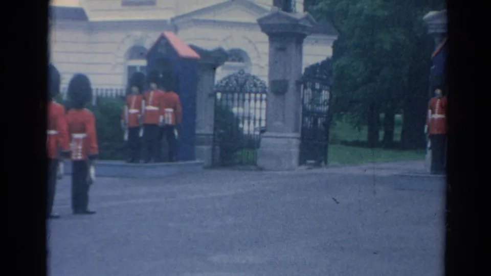 1964 London home-movie frame showing ceremonial guards near the Buckingham Palace entrance, with gates and stonework behind them.