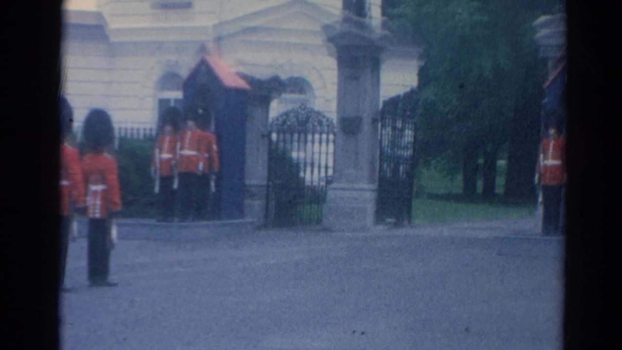 Guards in red tunics and tall black hats stand at the Buckingham Palace entrance in a soft 8mm London tourist view.