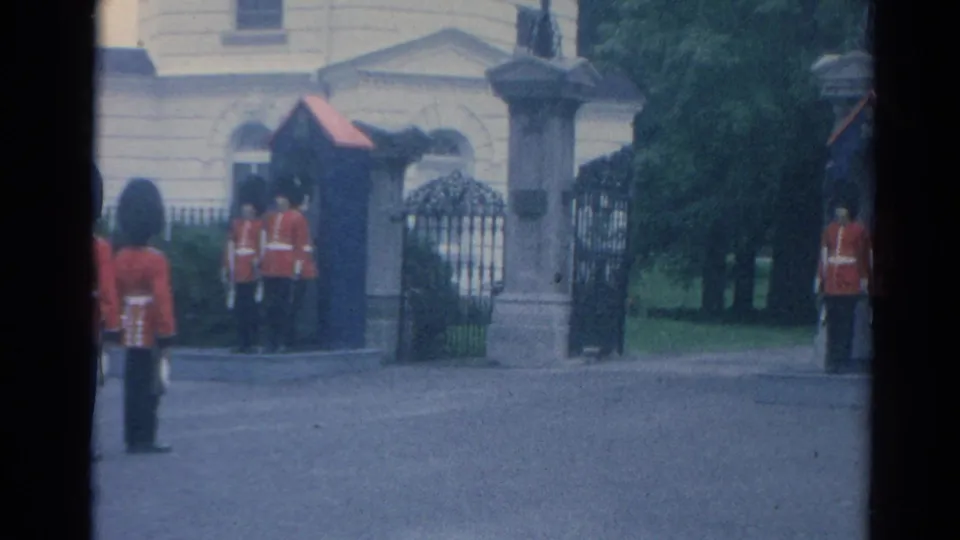 1964 Buckingham Palace entrance view with guards at attention, stone gateposts, iron gates, and trees in the background.