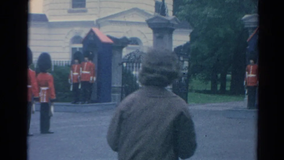 A passerby in a coat moves into the foreground of the 1964 London view while guards remain posted at Buckingham Palace.