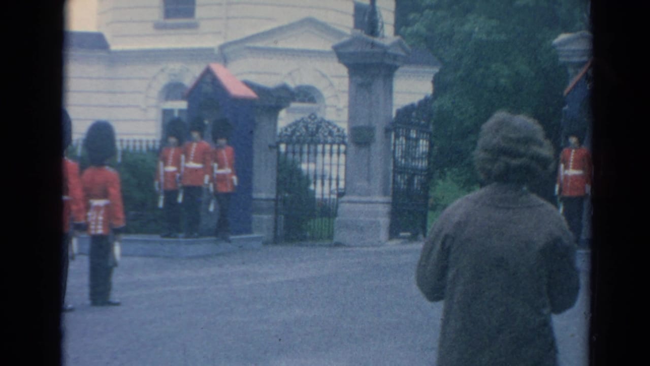 1964 London home-movie frame showing ceremonial guards by sentry boxes and iron gates, with a civilian observer in the foreground.