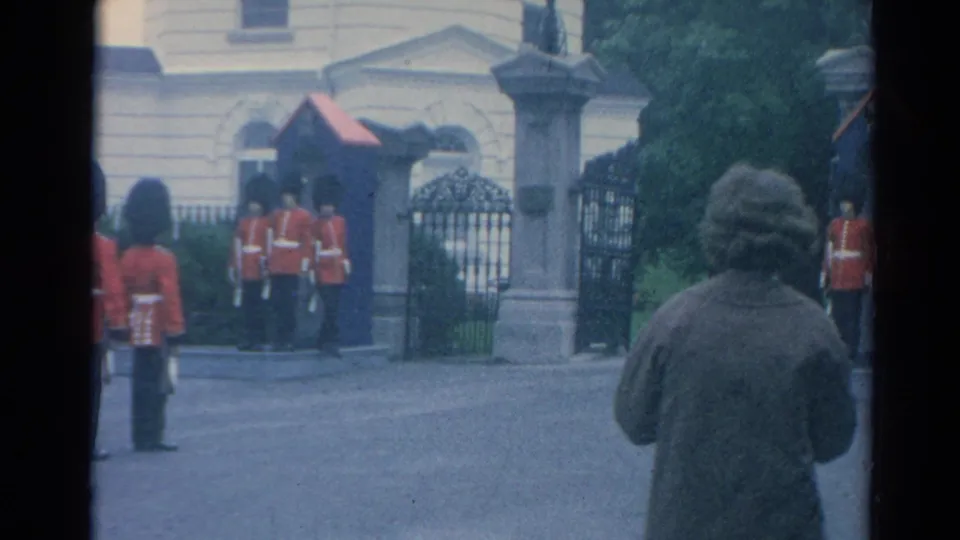 1964 London home-movie frame showing ceremonial guards by sentry boxes and iron gates, with a civilian observer in the foreground.
