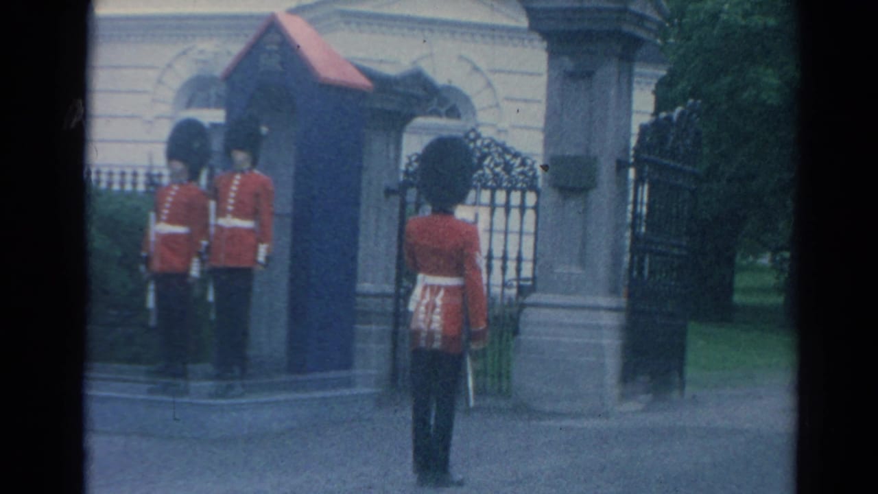 1964 London frame of a red-coated ceremonial guard standing near a sentry box, open iron gate, stone pillar, and greenery.