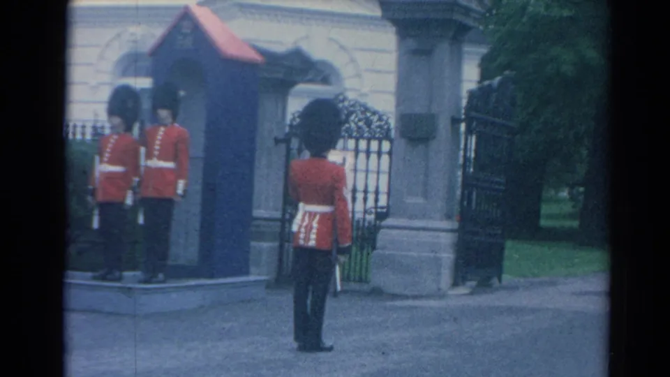 1964 London home-movie frame with red-coated guards positioned around a blue sentry box and iron gate during ceremony.