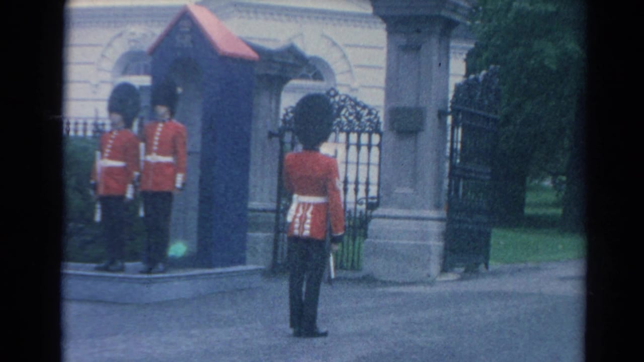 1964 London home-movie frame showing red-coated British guards near a blue ER sentry box, stone pillars, and wrought-iron gates.