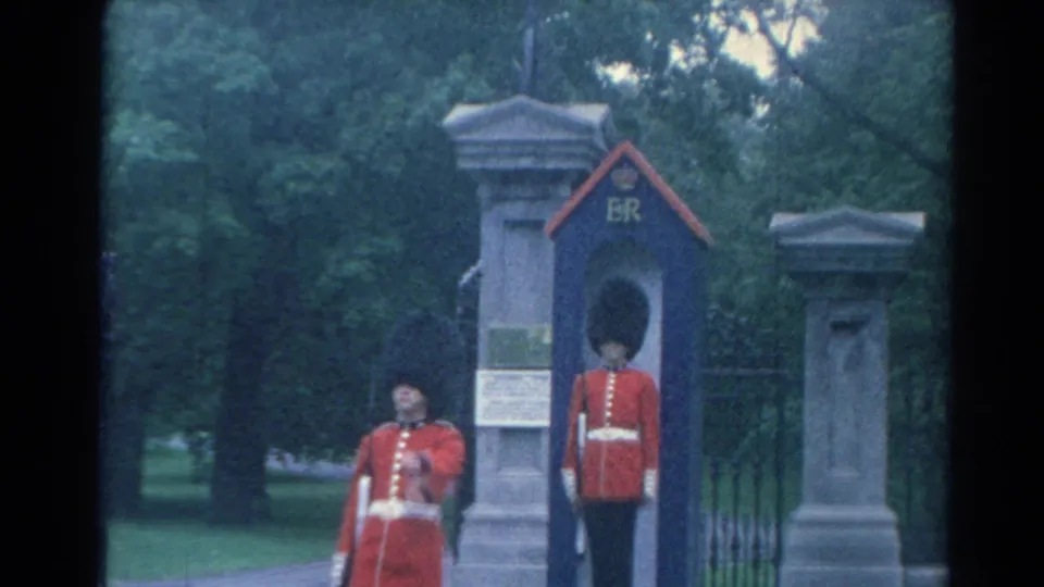 1964 London home-movie frame showing a guard at attention in an ER sentry box as another guard passes nearby.