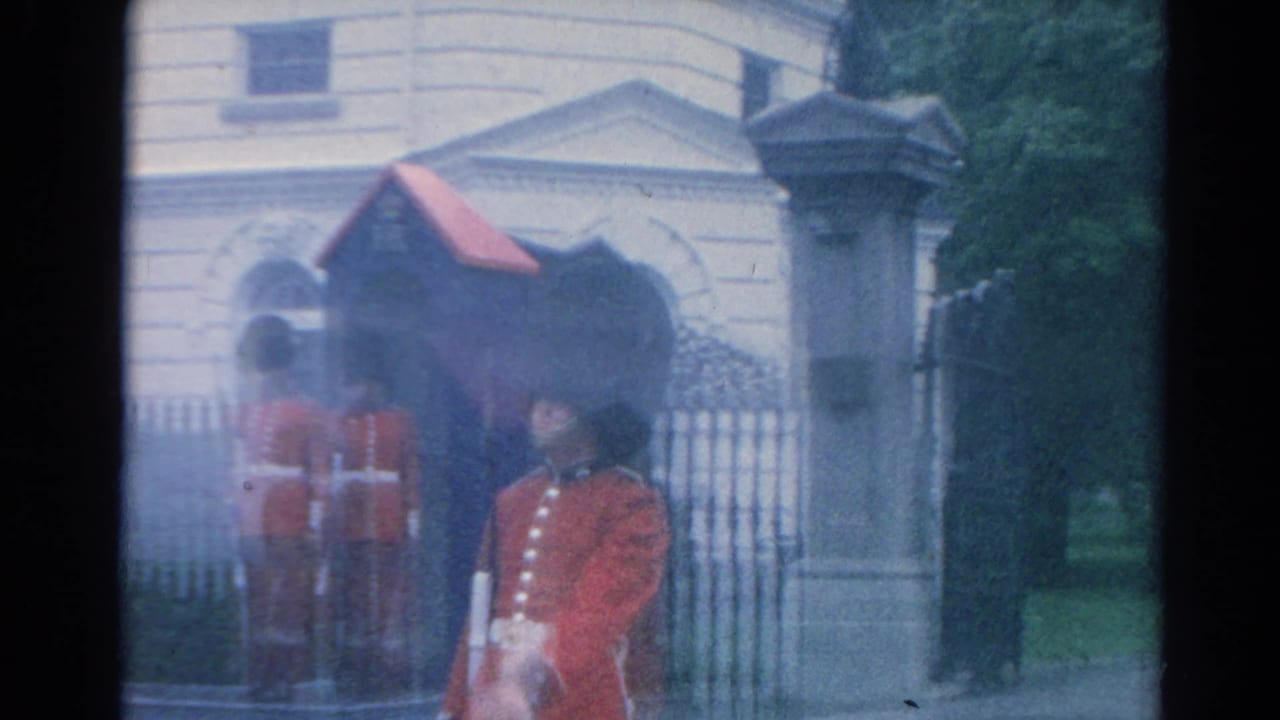 Several red-uniformed guards stand around a palace gate in London, with pale masonry and iron fencing behind them.