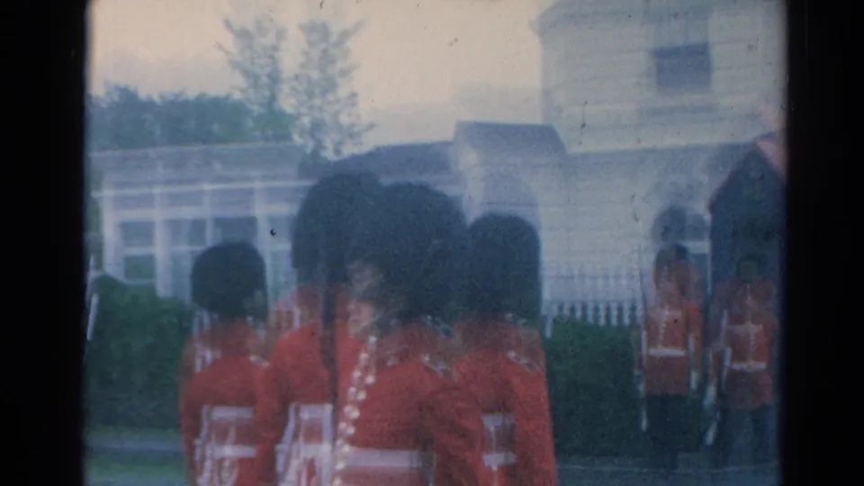 Red-coated guards are filmed from behind near the palace gate, with another guard standing by the entrance in soft 8mm focus.