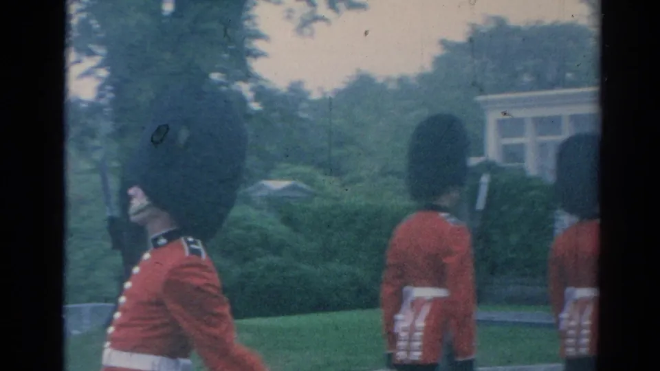 Red-uniformed guards march across the palace grounds in London, passing lawns, trees, and a pale structure in the background.