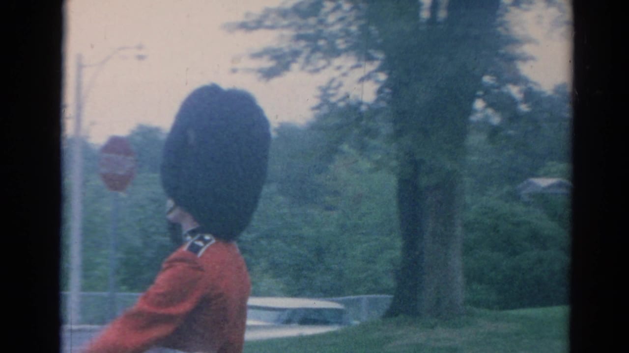 1964 London home-movie frame showing a British guard in red ceremonial dress and tall black hat near trees and roadside signage.