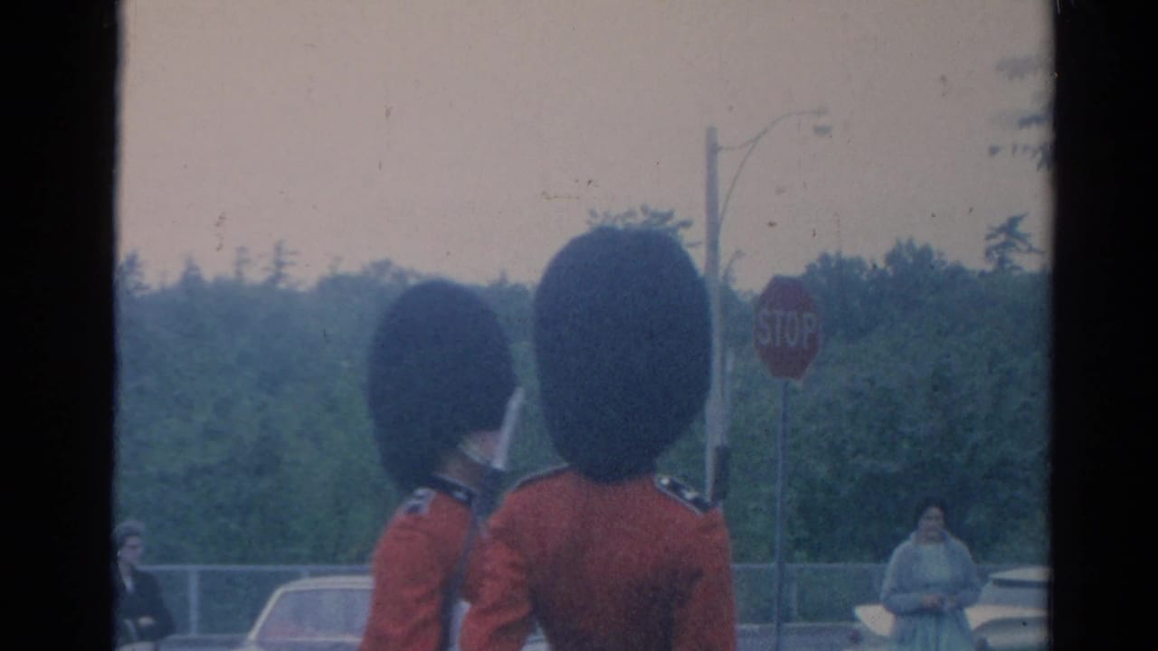 Two red-coated guards in tall black hats stand or march near a stop sign and spectators.