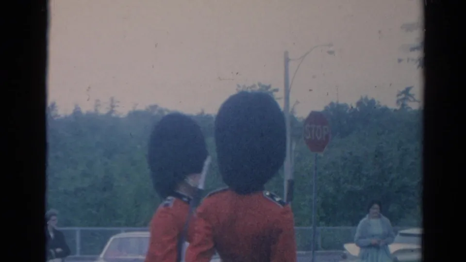 Two British guards in ceremonial red coats and tall black hats are filmed from behind in an outdoor London roadside setting.