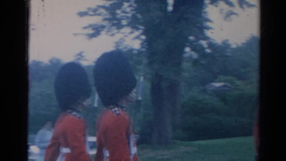1964 London frame with two ceremonial guards in red coats and tall black hats moving through a green roadside area.