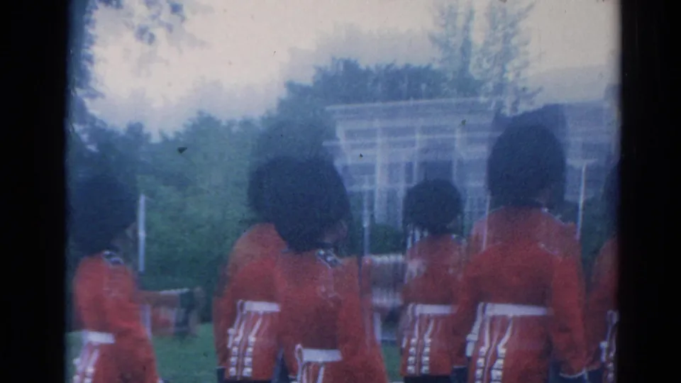 Several British guards in red ceremonial coats and tall black hats march away outdoors in 1964 London near railings and a pale building.