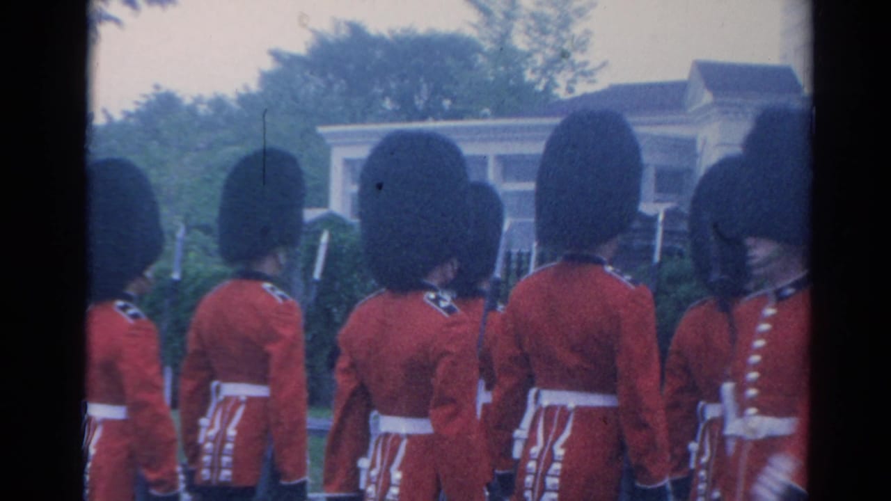 1964 London home-movie frame showing British ceremonial guards in red tunics marching in formation near a building and greenery.