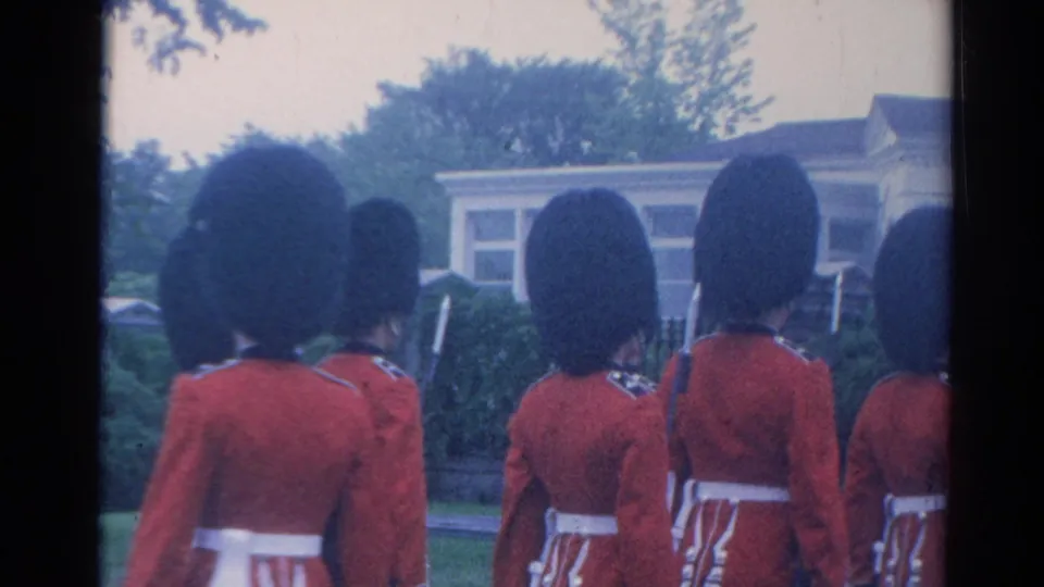 1964 London frame showing a rear view of ceremonial guards in red tunics and tall black hats moving together past trees and a building.