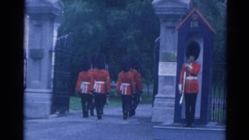 1964 London home-movie frame with guards marching through a stone gate as a sentry stands at attention beside an ER-marked box.