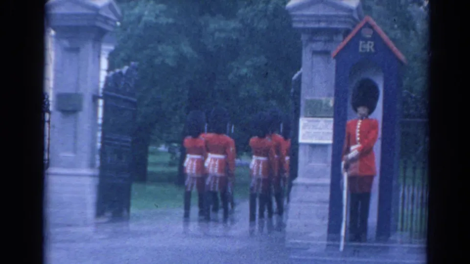 1964 London frame showing a ceremonial sentry beside an ER-marked guard box while guards recede through the gate into a tree-lined area.