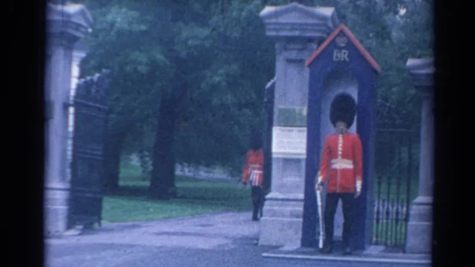 A remaining marching guard recedes along the path as the London sentry stands beside the blue box and stone gate on 8mm film.