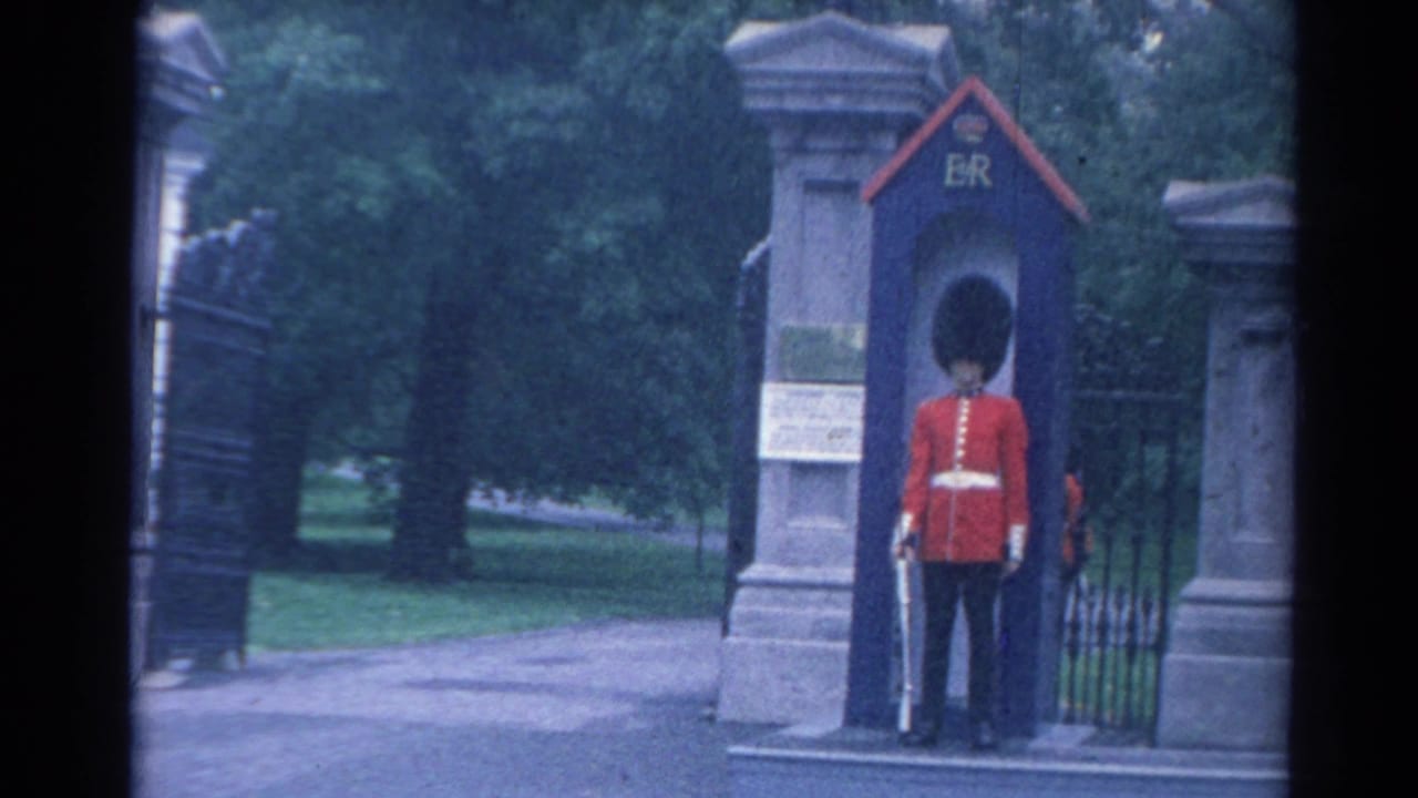 1964 London home-movie frame showing a guard in red uniform and tall black hat standing at attention in an ER-marked sentry box.