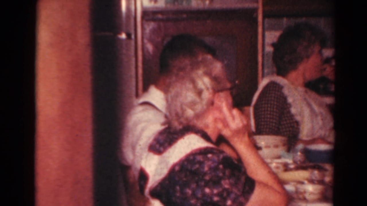 Adults sit close together at a dining table in a warmly lit 1960s home-movie kitchen interior.