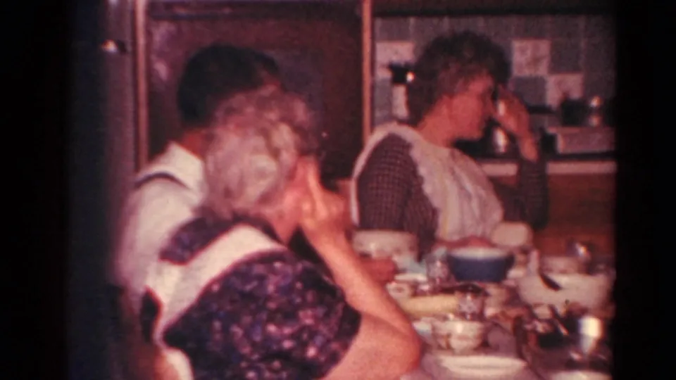 Home-movie frame of adults seated around a crowded dining table, with dishes and kitchen cabinets visible behind them.