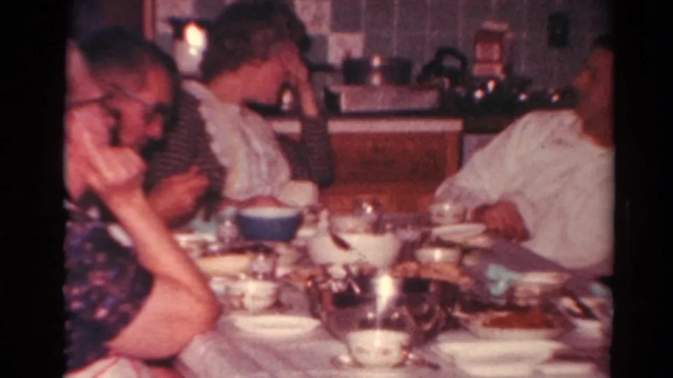 Adults sit around a kitchen dining table in 1964 home-movie footage, with tableware and a domestic counter in the background.