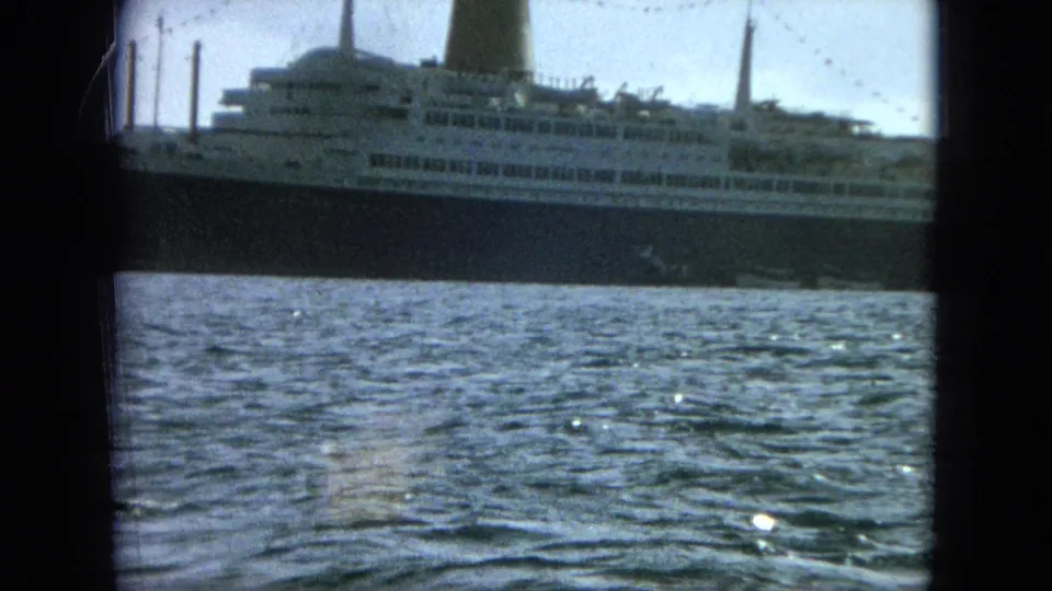 A closer 8mm frame shows the ship's midsection, white superstructure, dark hull, funnel, masts, and textured harbor water.