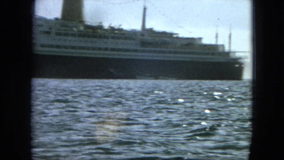 The vessel continues across the frame, with its long dark hull, white decks, funnel, stern area, masts, and rippled water visible.