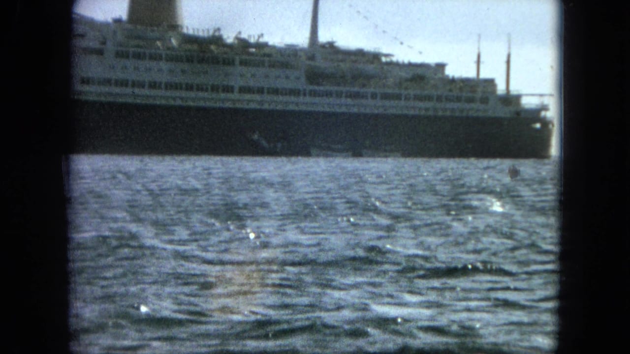 A multi-deck ocean liner is filmed from across the water, with visible grain, slight blur, and dark frame edges.