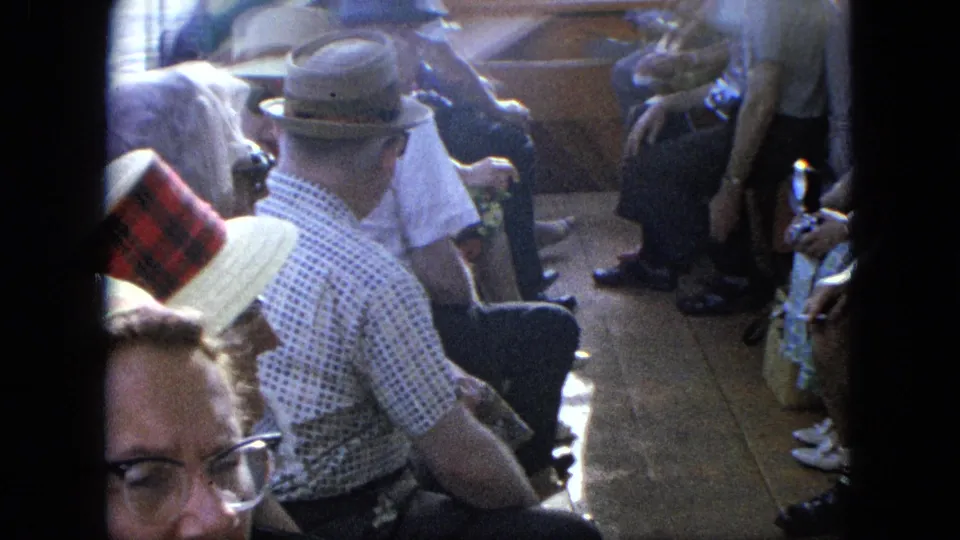 Older adults sit along a wooden boat deck in summer clothing, filmed candidly in grainy 8mm color.