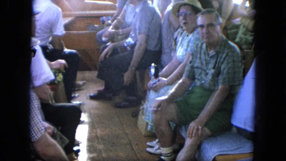 A group of older passengers in summer attire sits on a boat interior, with faces visible and some looking at the camera.