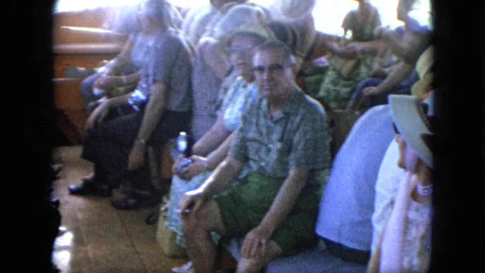 1964 home-movie frame of adults seated on a boat, including a man in sunglasses and green shorts facing the camera.