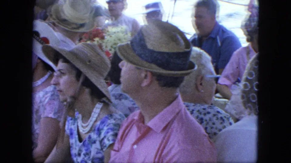 Crowded boat-deck view with adults in straw hats, a woman in a patterned dress, and water visible behind them.