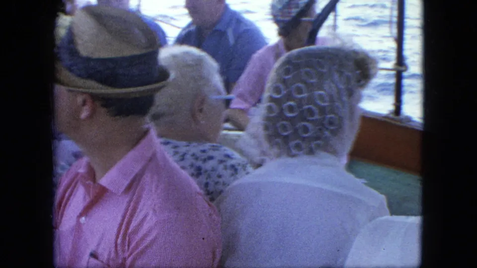 Home-movie frame from a boat ride showing passengers from behind and side angles, including hats and patterned head covering.