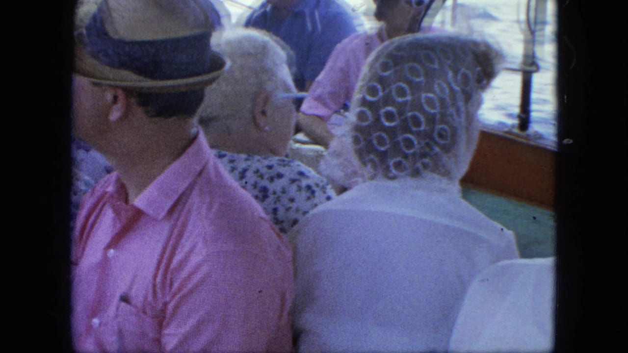 Adults seated on a ferry deck beside water, including a man in a pink shirt and fedora.