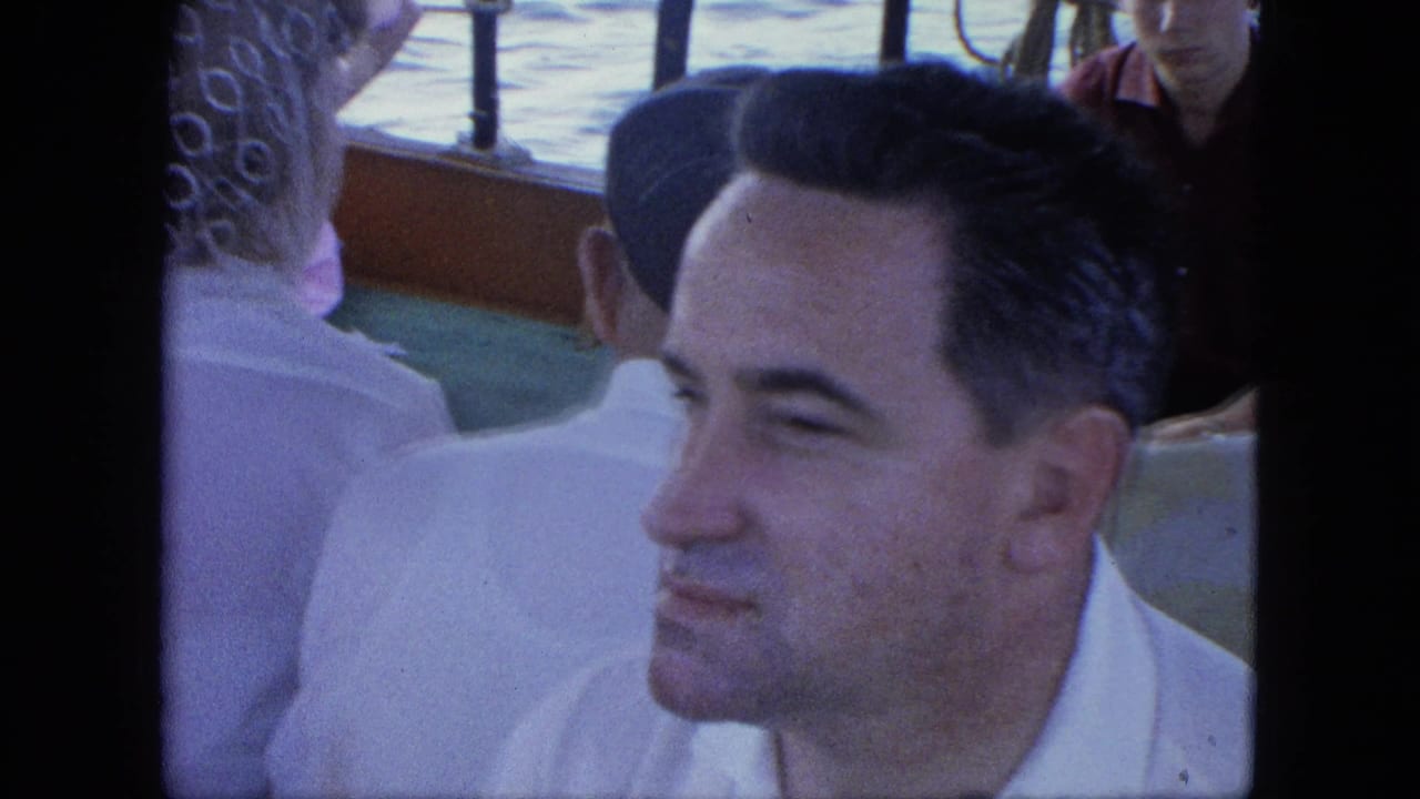 Adult man in a white shirt sits among crowded ferry passengers, with railings and water visible behind him.