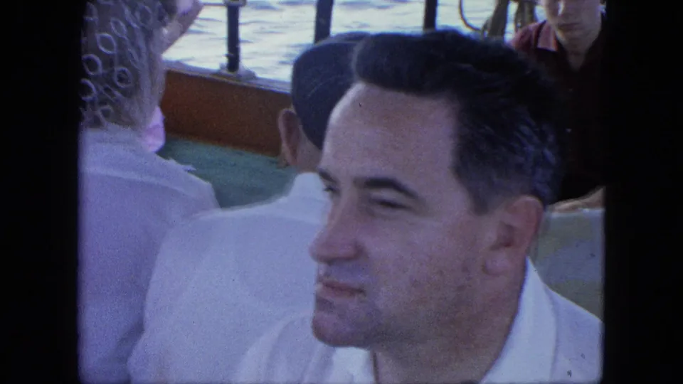 Adult man in a white shirt sits among crowded ferry passengers, with railings and water visible behind him.