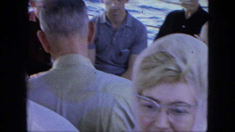 Passengers sit close together on the ferry deck, including a foreground woman with glasses and a man seen from behind.