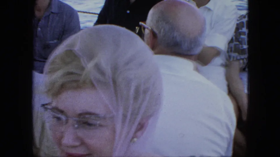 A woman wearing a light headscarf and glasses sits among adult ferry passengers, with water and railings behind the group.