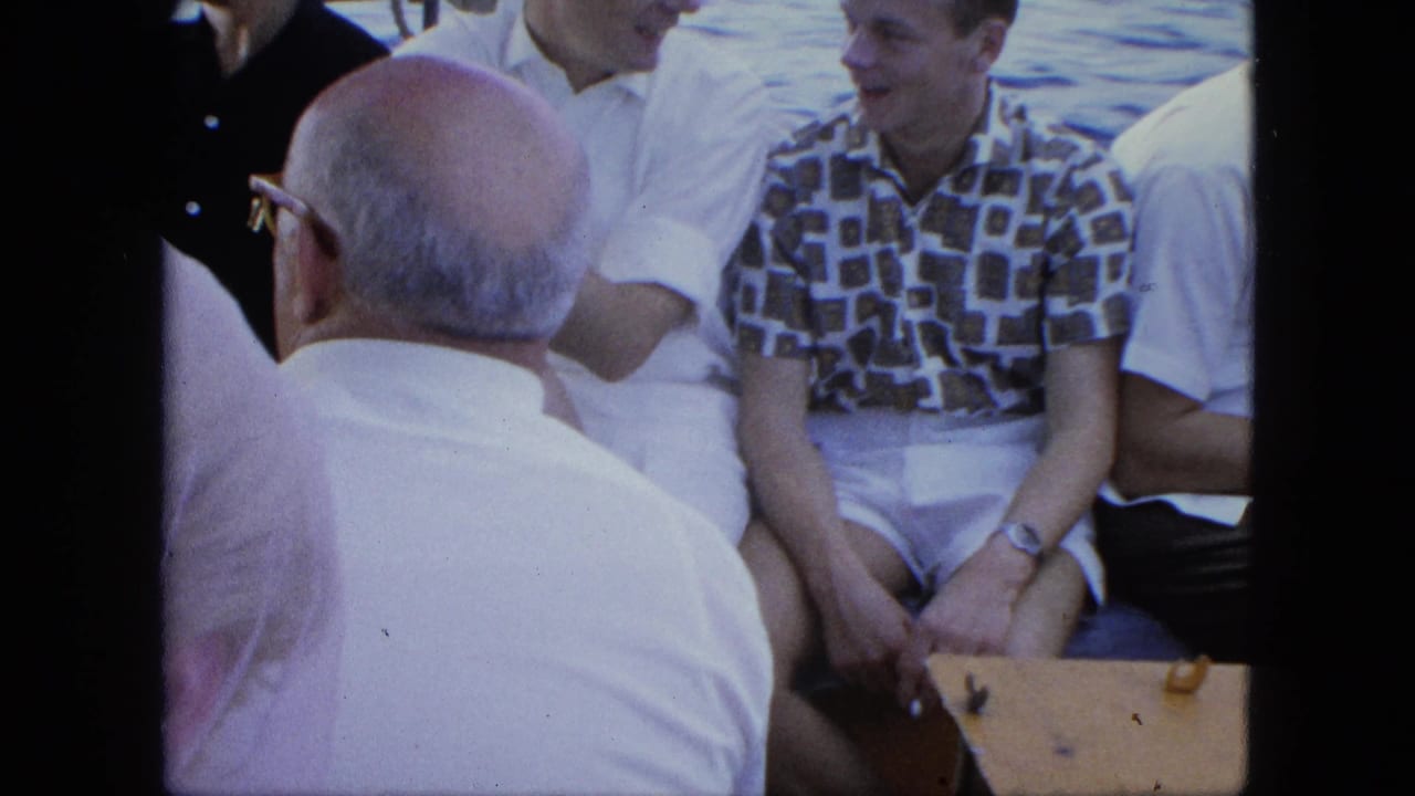 Adults sit close together on a boat, with a bald man seen from behind and passengers in light summer clothing.
