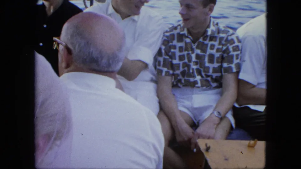 1964 8mm home-movie frame of adults seated on a small boat, including a man seen from behind and passengers in white and patterned clothing.